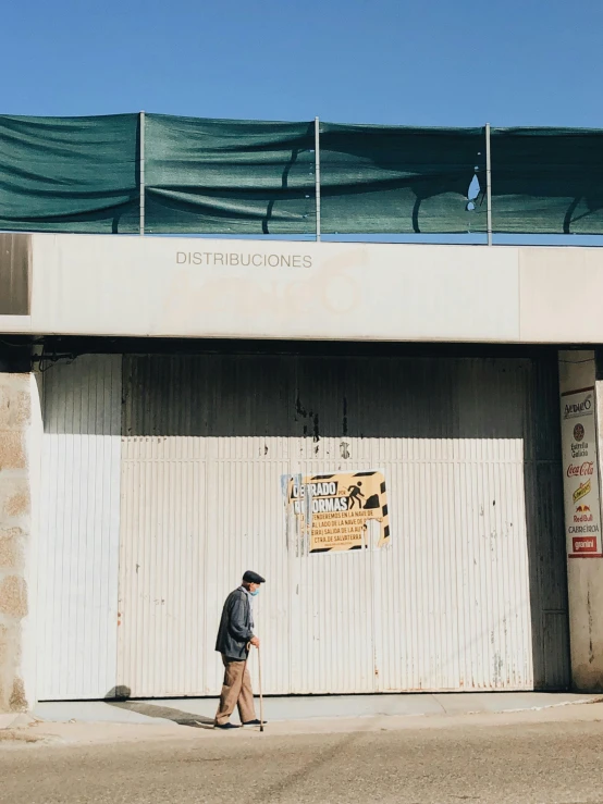 a man walking down a sidewalk in front of a building, an album cover, by Alejandro Obregón, unsplash, entrance to abandoned mine, 😭 🤮 💕 🎀, refrigerated storage facility, los carpinteros