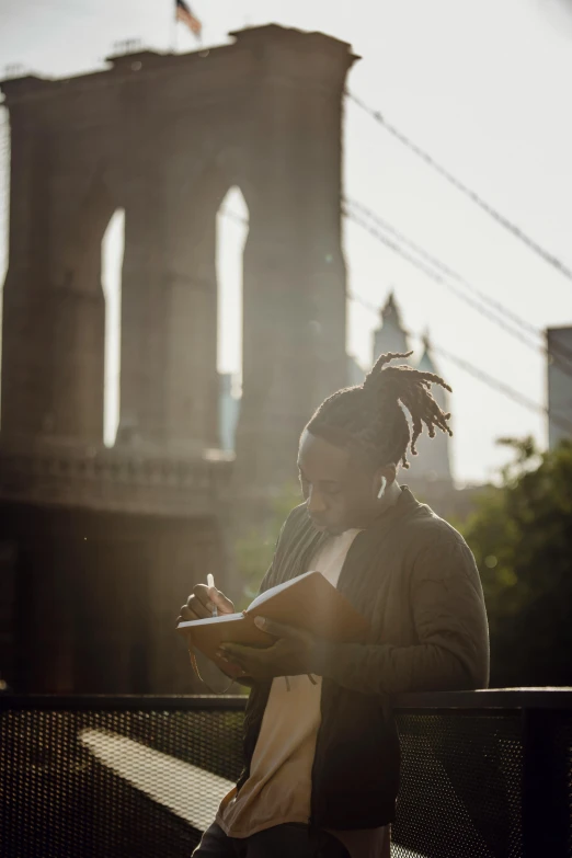 a person sitting on a bench in front of a bridge, academic art, holding notebook, brooklyn, morning light, dark skinned