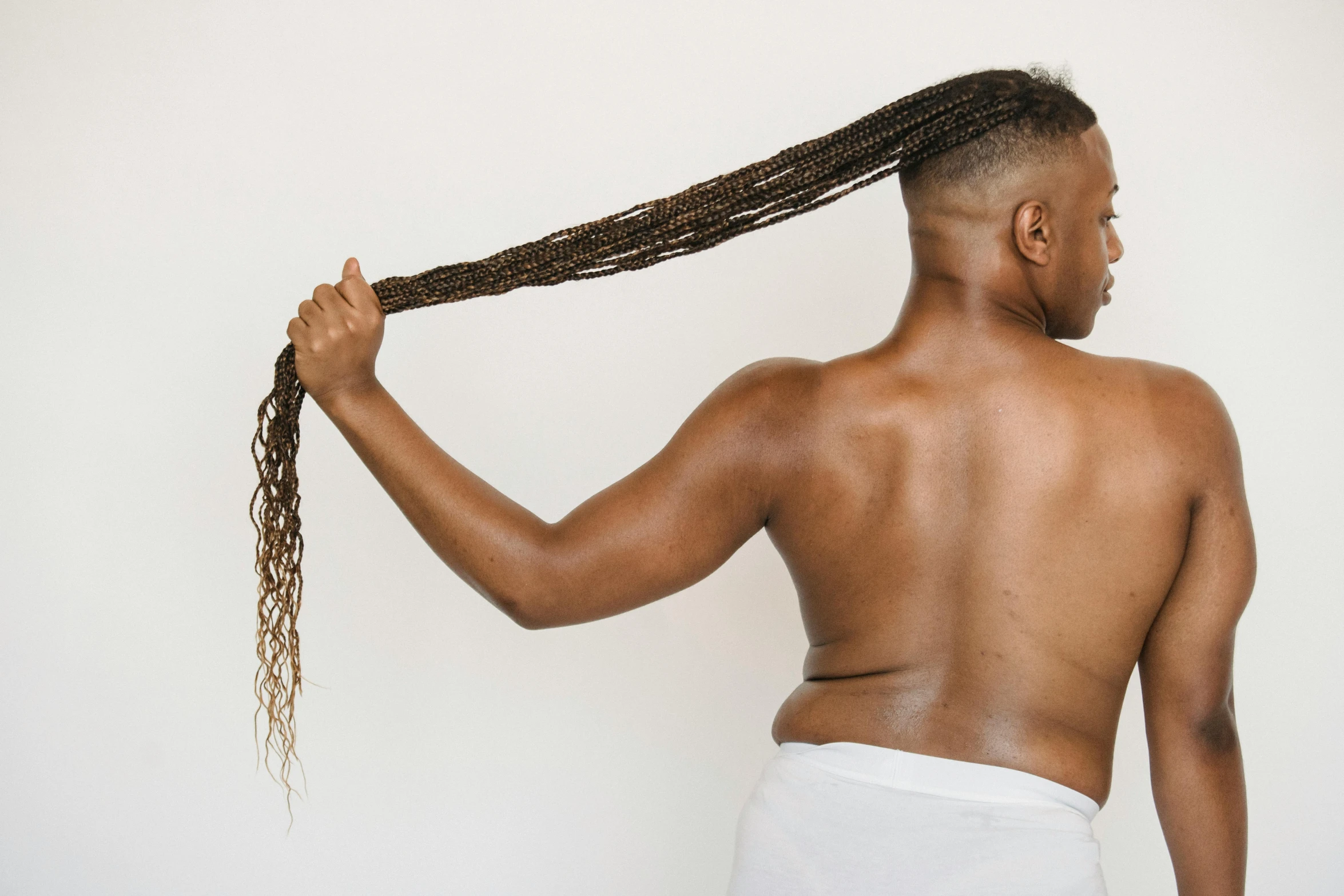 a man with long hair standing in front of a white wall, trending on pexels, afrofuturism, waist reaching ponytail, disrobed, complex braided hair, an obese