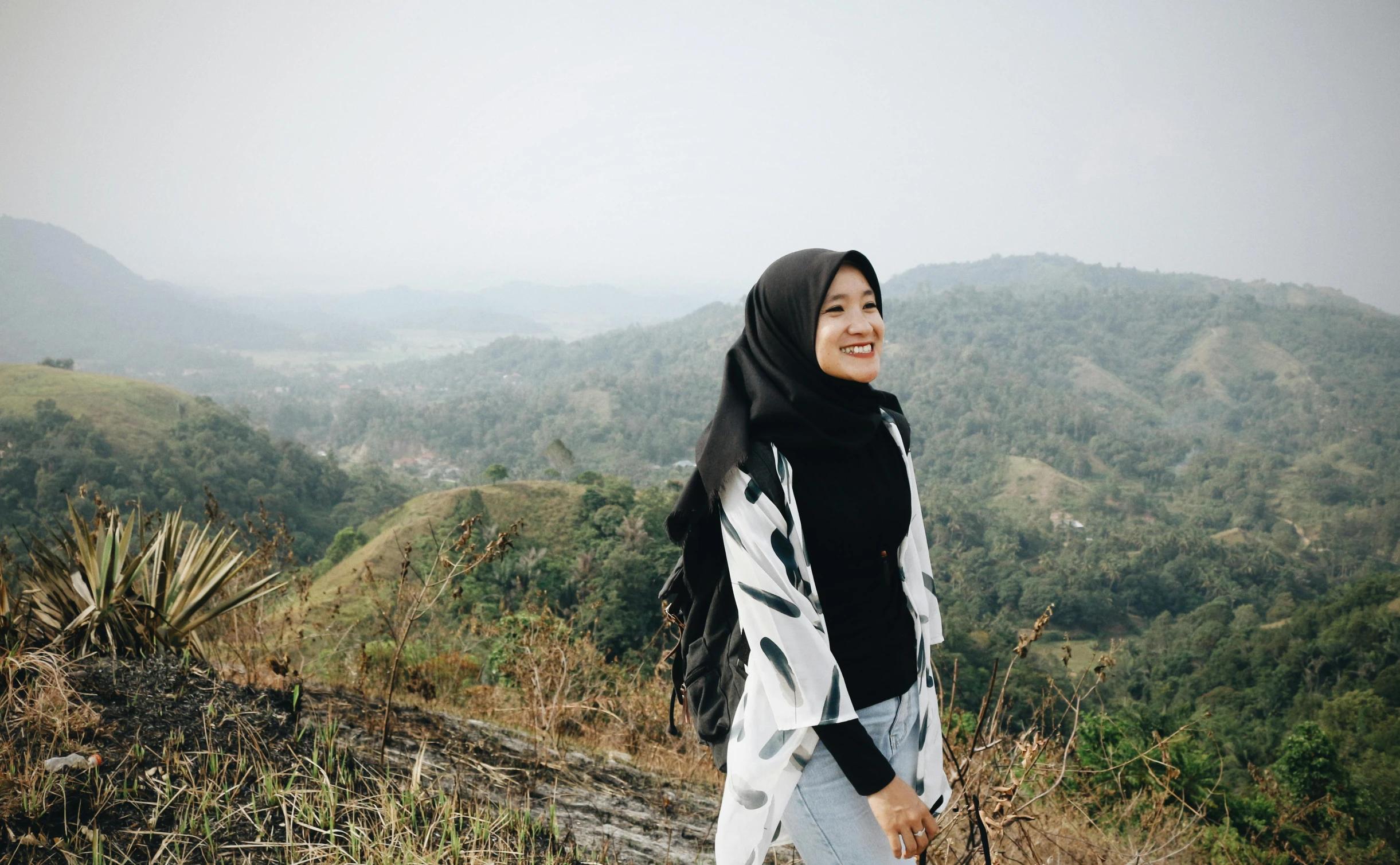 a woman standing on top of a lush green hillside, inspired by Nazmi Ziya Güran, pexels contest winner, hurufiyya, wrapped in a black scarf, casual modern clothing, with a long white, malaysian