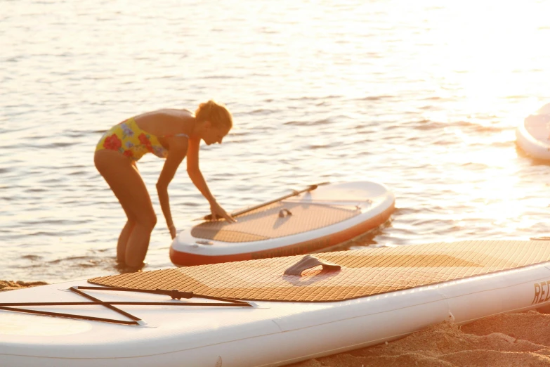 a woman standing on top of a surfboard on a beach, boats in the water, golden sunlight, maintenance, inflatable