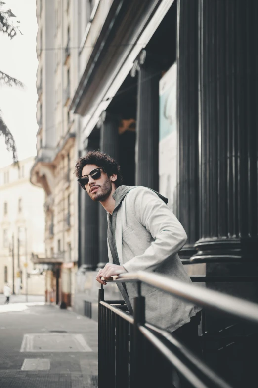 a man leaning on a railing on a city street, pexels contest winner, beige hoodie, daniel ricciardo, in suit with black glasses, hozier