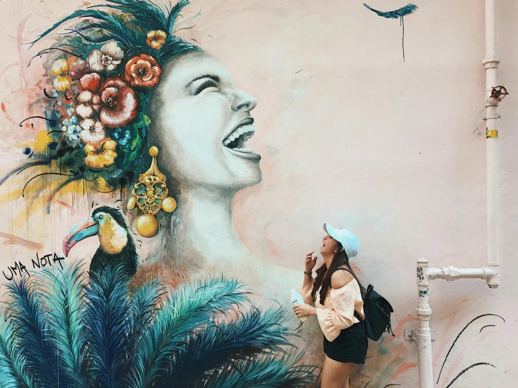 a woman standing in front of a wall with a painting on it, pexels contest winner, street art, feathered headdress, happy girl, flora and fauna, beautiful painting of a tall