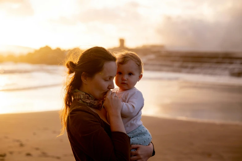 a woman holding a baby on a beach, by Matt Stewart, pexels contest winner, portrait soft light, morning glow, high quality upload, gold