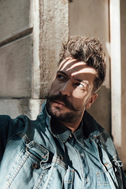 a man in a denim jacket leaning against a wall, an album cover, inspired by Germán Londoño, trending on pexels, renaissance, he got a big french musctache, guy fieri, headshot profile picture, pompadour