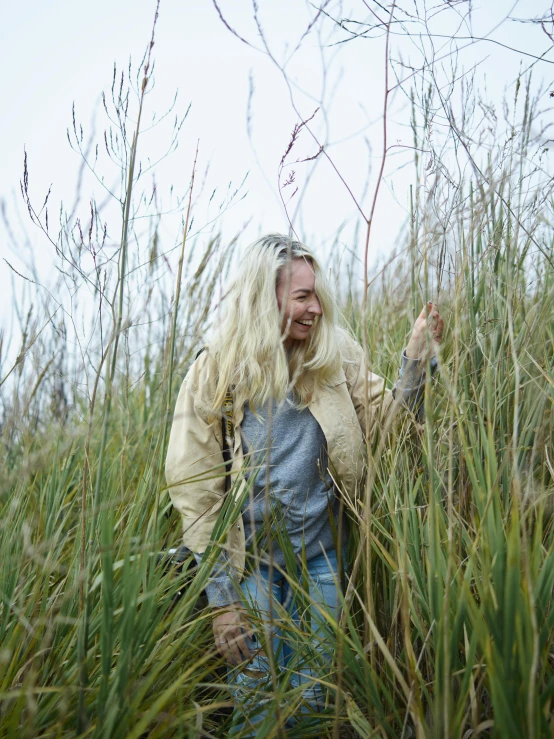 a woman standing in a field of tall grass, a picture, by Caroline Mytinger, land art, wearing farm clothes, blonde swedish woman, smiling playfully, on the coast