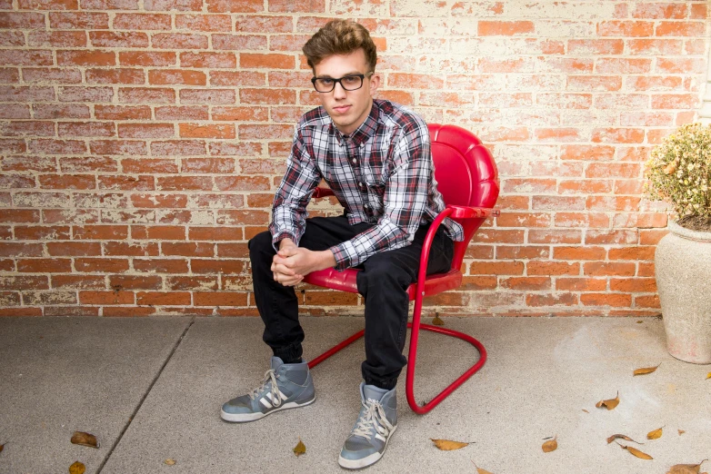 a man sitting on a red chair in front of a brick wall, an album cover, inspired by Max Gubler, pexels, wearing glasses, teenage boy, casual pose, red shoes