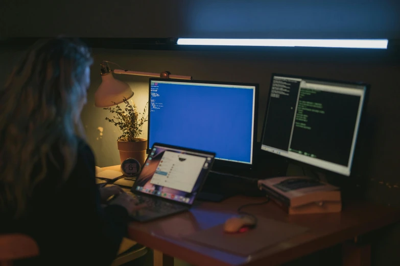 a woman sitting at a desk in front of two computer monitors, by Adam Marczyński, pexels, night time, a python programmer's despair, avatar image, bottom angle