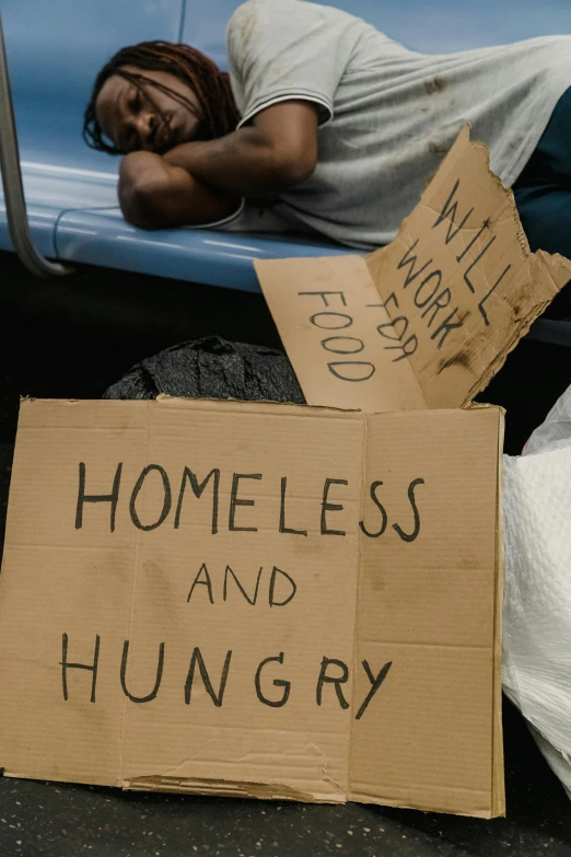 a man sleeping on the ground next to a sign that says homeless and hungry, a photo, stacked image, cardboard, subway, (digital art)