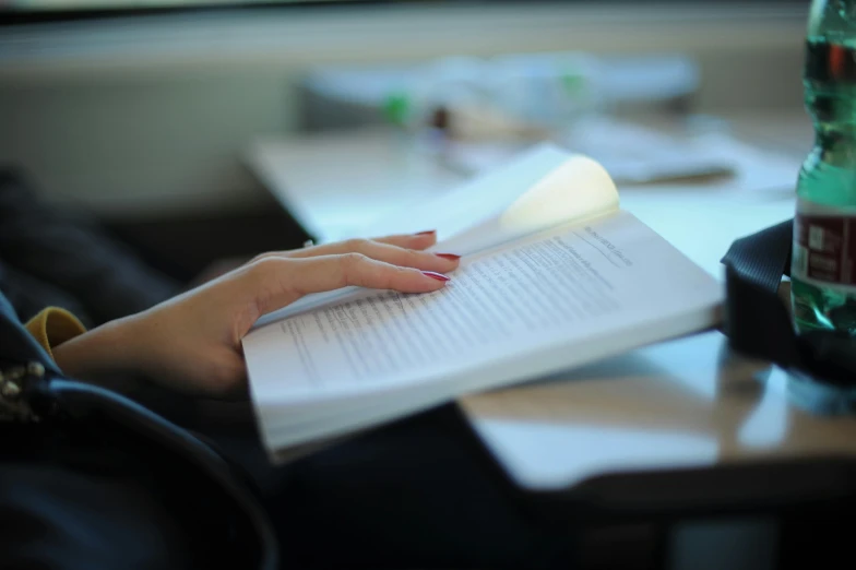 a woman sitting at a table reading a book, by Jessie Algie, pexels contest winner, light emitting from fingertips, sitting in the classroom, soft light from the side, paper