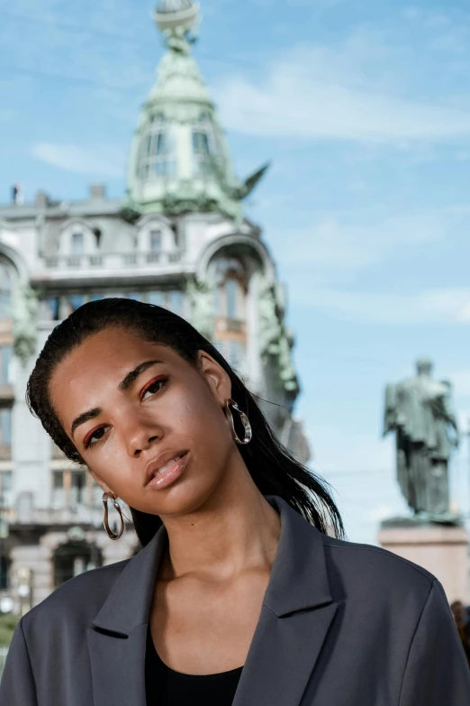 a woman standing in front of a building with a clock tower in the background, by Mathias Kollros, trending on pexels, baroque, brown skin. light makeup, helsinki, zoë kravitz, serious business