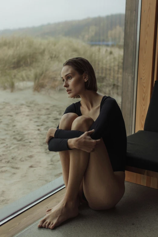 a woman sitting on the floor in front of a window, a picture, by Anna Boch, unsplash contest winner, natalie portman, standing at the beach, wearing a black bodysuit, sad man