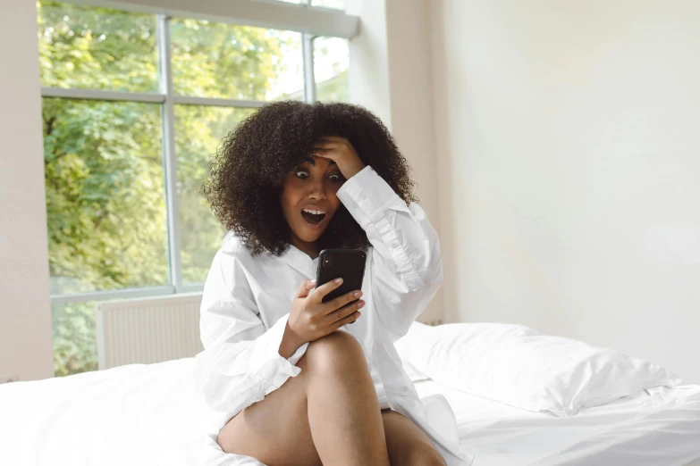 a woman sitting on top of a bed holding a cell phone, trending on pexels, happening, curly afro, surprised, white sheets, white dress shirt