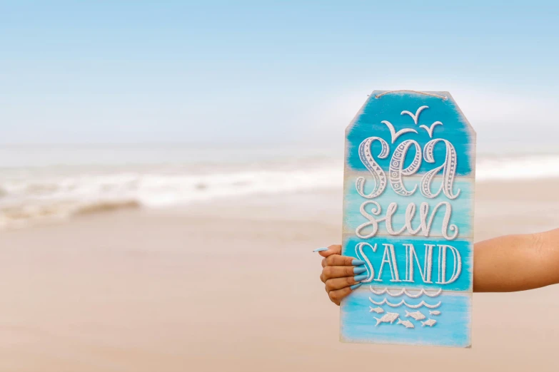 a person holding up a sign on the beach, pexels contest winner, handpainted, svg sticker art, blue sand, decoration