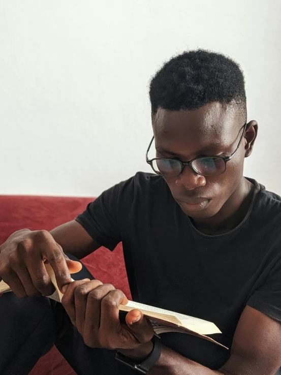 a man sitting on a couch reading a book, by Chinwe Chukwuogo-Roy, pexels contest winner, afrofuturism, professional woodcarving, in square-rimmed glasses, male teenager, holding scimitar made of bone