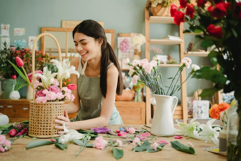 a woman sitting at a table with a basket of flowers, making the best smug smile, crafting, photo for a store, fine detailing