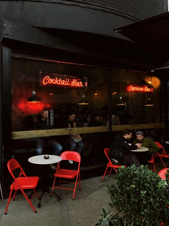 a group of people sitting at tables outside of a restaurant, red and obsidian neon, cosy vibes, thumbnail, s baraldi