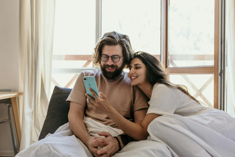 a man and woman sitting on a bed looking at a cell phone, trending on pexels, bushy beard, mia khalifa, happy couple, australian