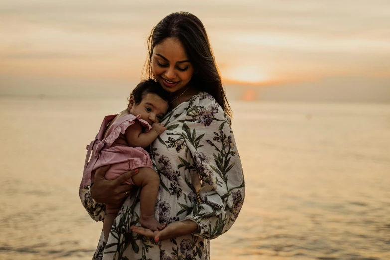 a woman holding a baby near a body of water, pink golden hour, an olive skinned, wearing pink floral chiton, full frame image