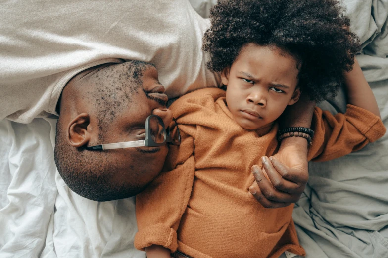 a man and a child laying on a bed, pexels contest winner, brown skinned, scowl, curls on top of his head, looking her shoulder