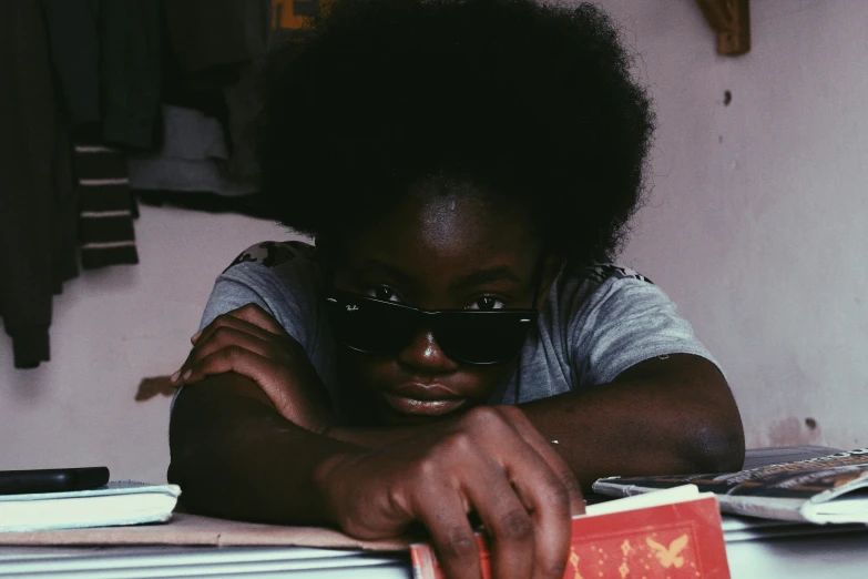 a person sitting at a table with a book, pexels contest winner, afrofuturism, wearing shades, black teenage girl, close-up portrait film still, old school
