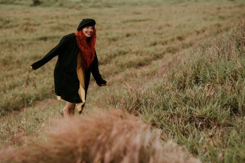 a woman walking across a grass covered field, a portrait, by Jessie Algie, pexels contest winner, happening, black and orange coat, happy friend, wild ginger hair, smiling playfully