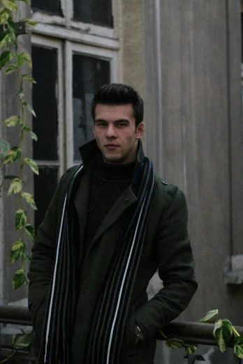 a man standing in front of a building wearing a scarf, dark green tones, young spanish man, wearing hunter coat, liam