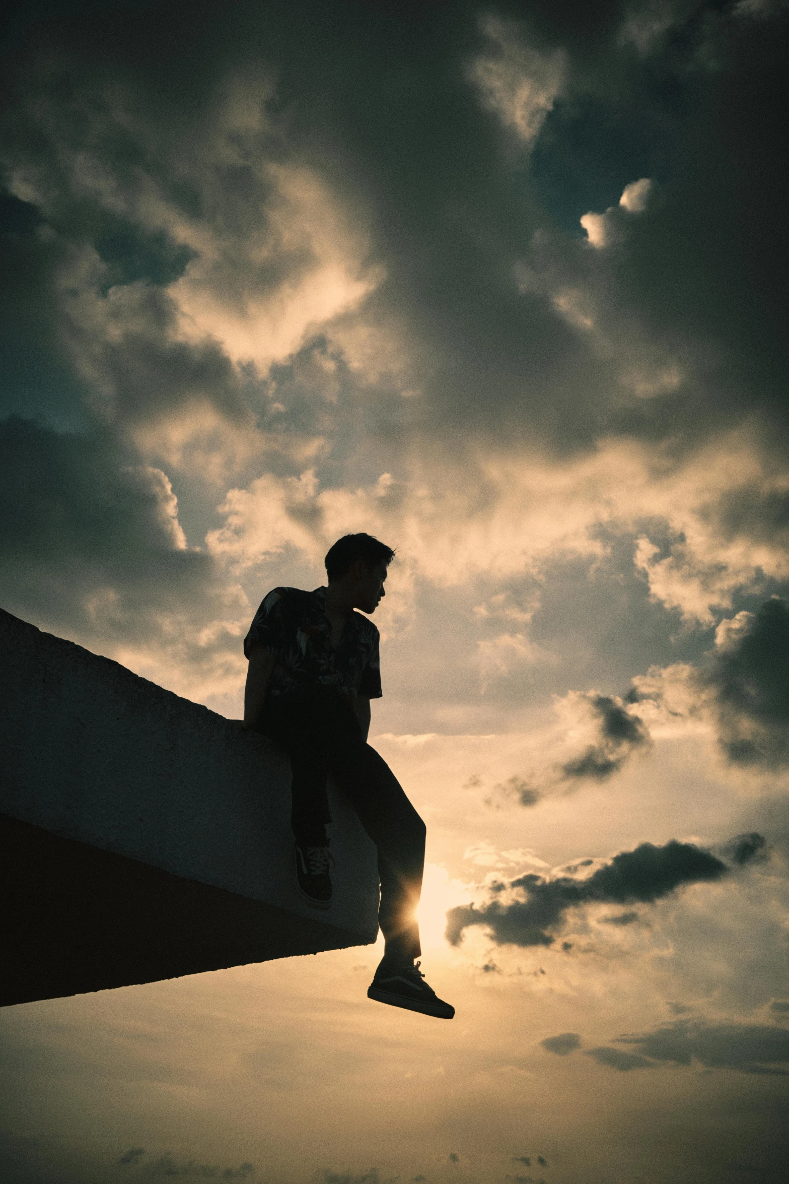 a man sitting on top of a building under a cloudy sky, a picture, trending on pexels, sunset lighting ominous shadows, teenage boy, sittin