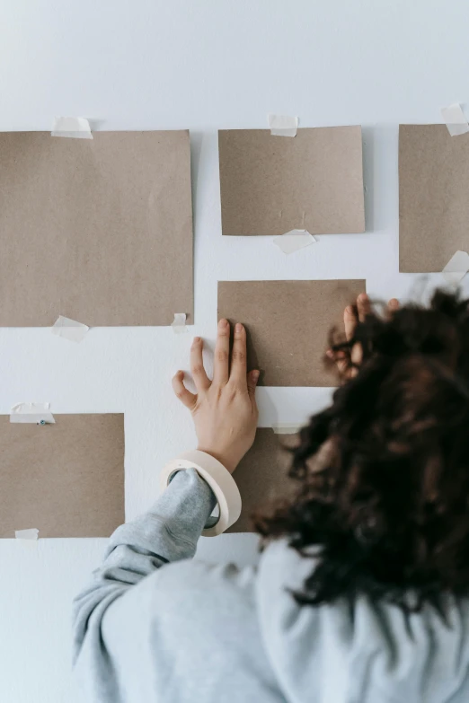 a woman standing in front of a wall covered in pieces of paper, pexels contest winner, visual art, square shapes, brown paper, inspect in inventory image, programming