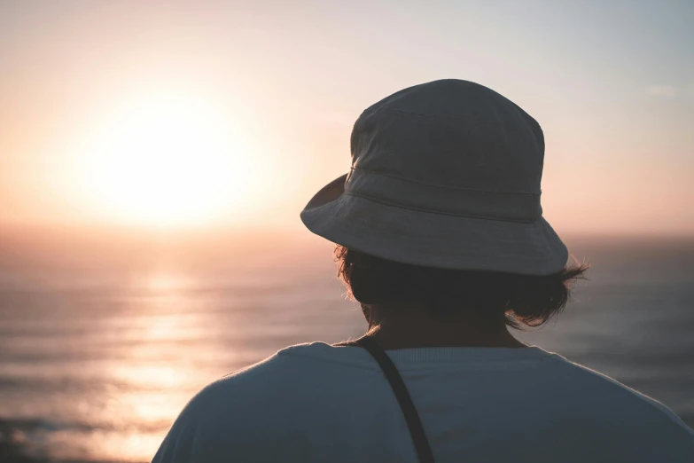 a person wearing a hat looking out at the ocean, trending on pexels, soft sunset lighting, bucket hat, in a sunset haze, manly