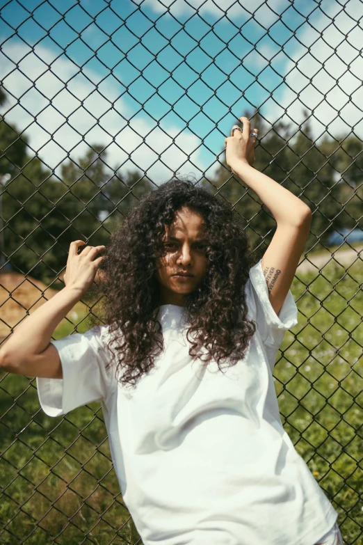 a woman leaning against a chain link fence, an album cover, pexels contest winner, dressed in a white t-shirt, curly dark hair, trending on vsco, asher duran