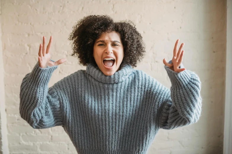 a woman standing in front of a brick wall with her hands in the air, trending on pexels, antipodeans, screaming in pain, blue turtleneck, curly haired, wearing an oversized sweater