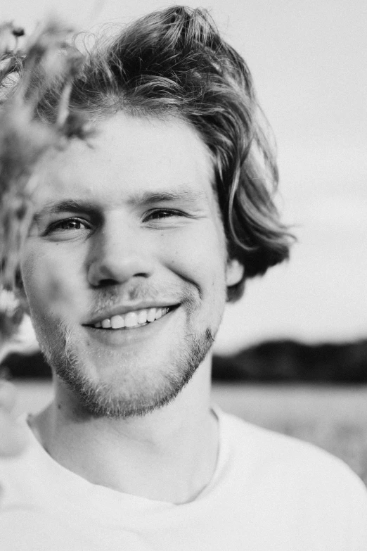 a black and white photo of a man smiling, by Jacob Toorenvliet, unsplash, happening, yung lean, hr ginger, shot at golden hour, his hair is messy and unkempt
