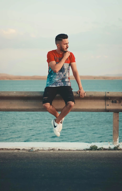 a man sitting on a railing next to the ocean, a picture, trending on pexels, patterned clothing, wearing an orange t shirt, dmitry prozorov style, gang clothing