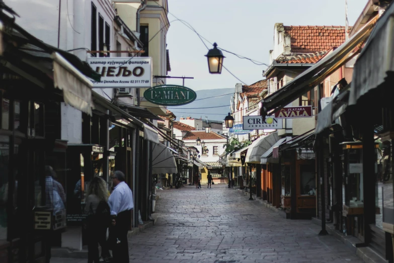 a couple of people that are walking down a street, by Matija Jama, pexels contest winner, renaissance, bosnian, lots of signs and shops, ground level view of soviet town, quiet street