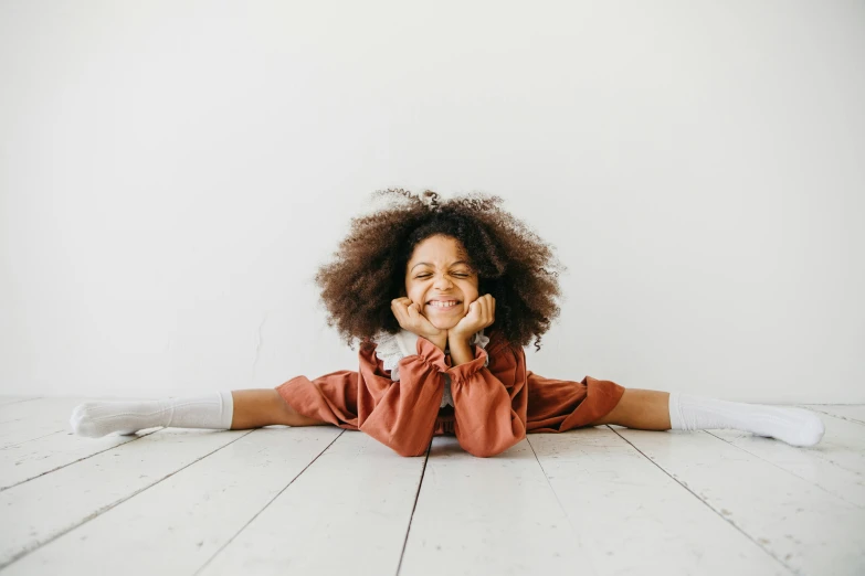 a little girl laying on the floor with her hands on her face, pexels contest winner, big afro, doing splits and stretching, smiling woman, sitting in an empty white room