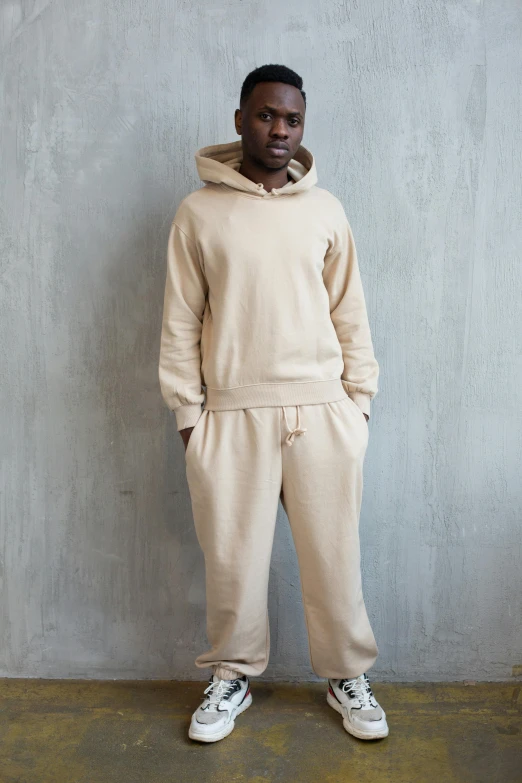 a man standing in front of a concrete wall, beige hoodie, wearing a hoodie and sweatpants, on display, light-brown skin