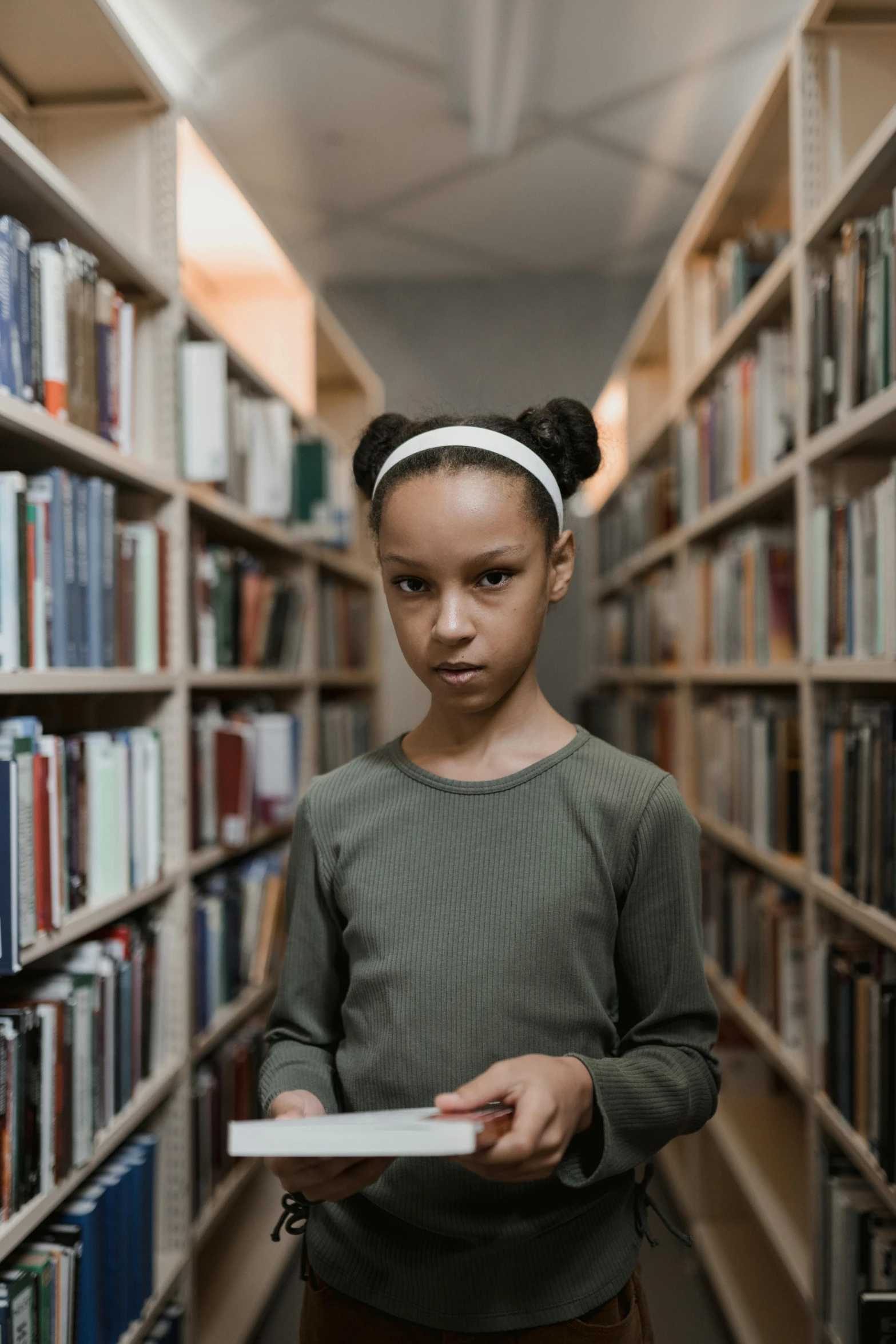 a young girl holding a book in a library, pexels contest winner, modernism, looking serious, brown skinned, standing, child hybrid