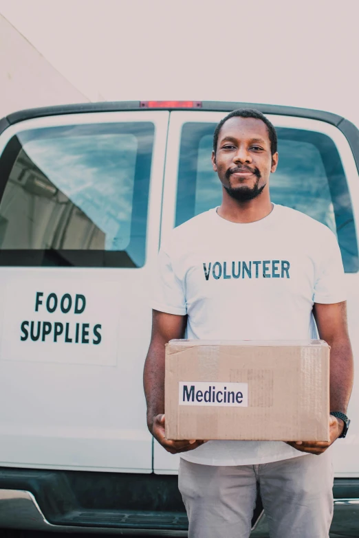 a man holding a box in front of a van, by Francis Helps, medical supplies, food, backdrop, portrait image