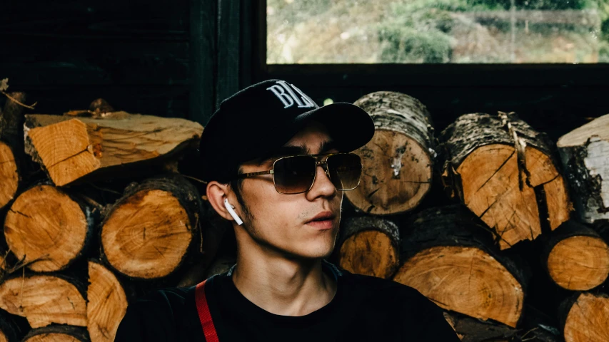 a man standing in front of a pile of logs, an album cover, inspired by Zhu Da, trending on pexels, earbuds jewelry, wearing sunglasses and a cap, headshot profile picture, dslr 5 0 mm portrait photo