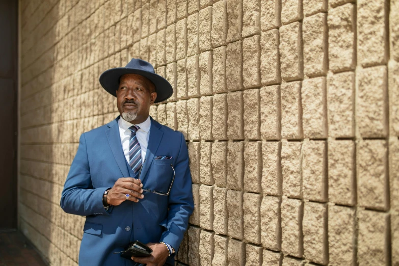 a man in a blue suit and hat leaning against a wall, mongezi ncaphayi, still image from tv series, shot on sony a 7, ap news