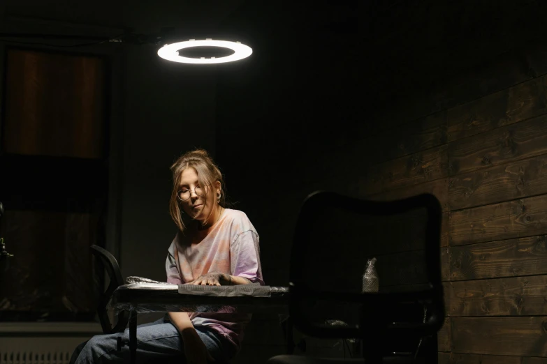 a woman sitting at a table in a dark room, bright halo, ring light, poor fluorescent lighting, sydney sweeney