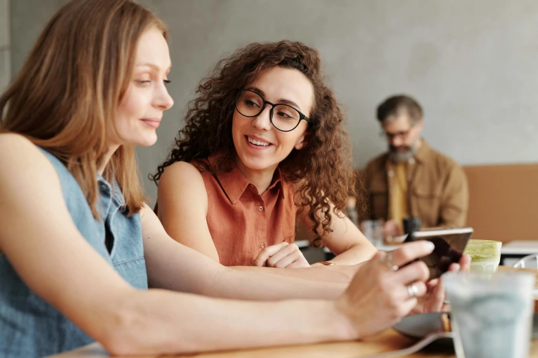 two women sitting at a table looking at a cell phone, trending on pexels, avatar image, schools, brown, high resolution image