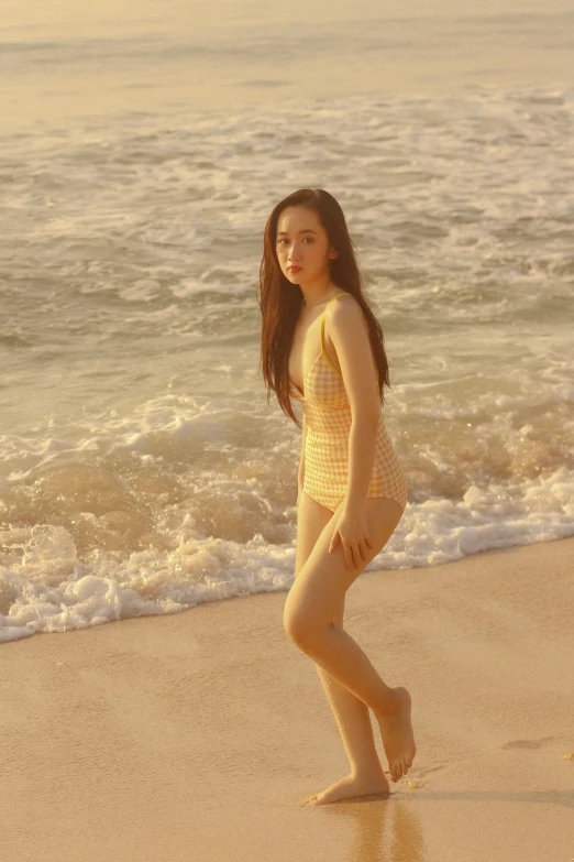 a woman standing on a beach next to the ocean, an album cover, by Han Gan, dilraba dilmurat, in a gold one piece swimsuit, taken at golden hour, gif