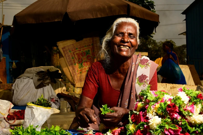 a woman sitting in front of a bunch of flowers, pexels contest winner, on an indian street, elder, thumbnail, smiling