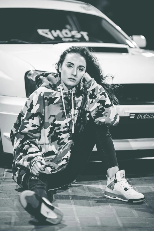 a woman sitting on the ground next to a car, a black and white photo, trending on pexels, antipodeans, wearing camo, mac miller, confident stance, wearing a hoody