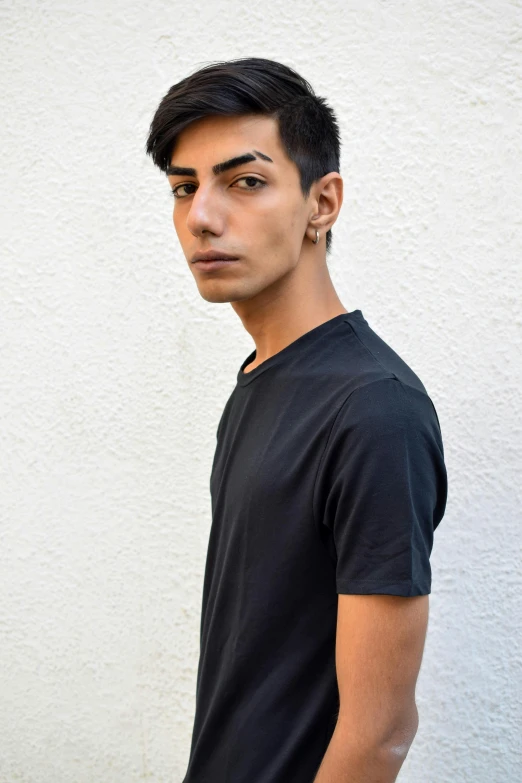 a young man standing in front of a white wall, an album cover, inspired by Nadim Karam, pexels contest winner, wearing a black t-shirt, an epic non - binary model, profile image, middle eastern