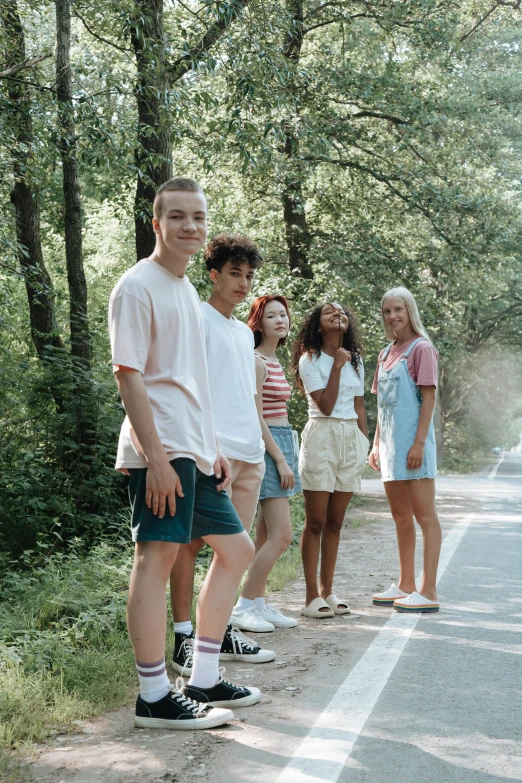 a group of people standing on the side of a road, trending on pexels, tan skin a tee shirt and shorts, male teenager, woodlands, lush surroundings