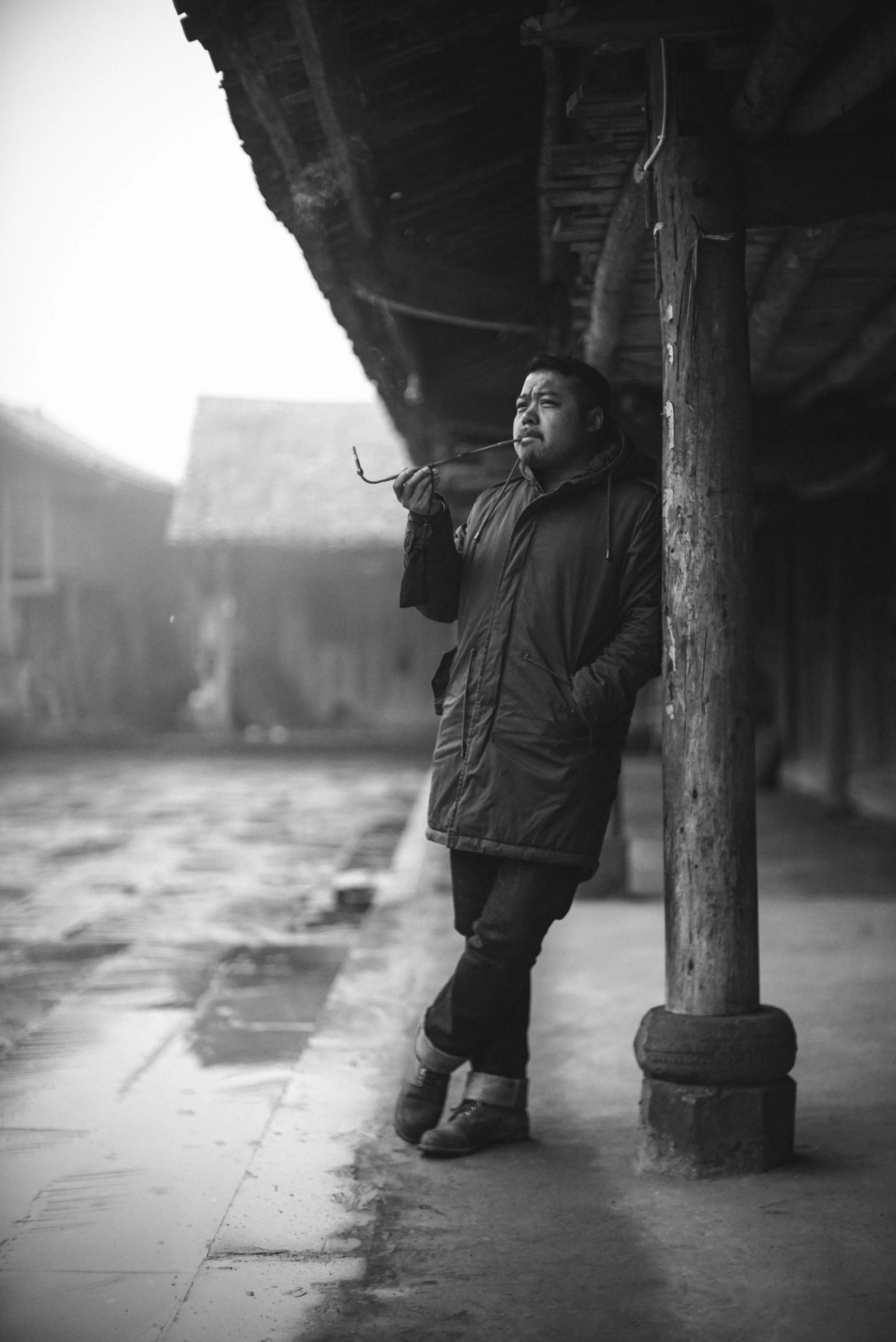 a black and white photo of a man leaning against a pole, inspired by Zhang Zongcang, pexels contest winner, with village, me, chilly, with cigar