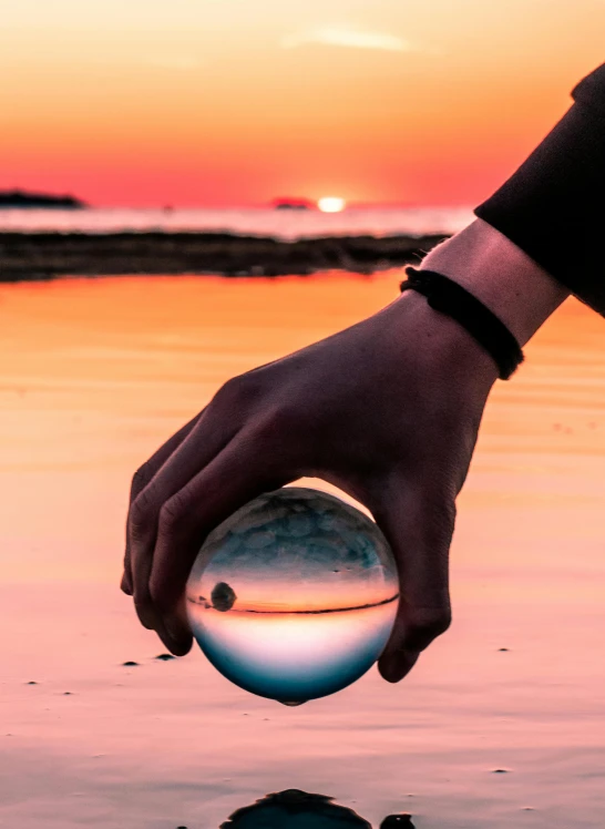 a person holding a glass ball over a body of water, during sunset